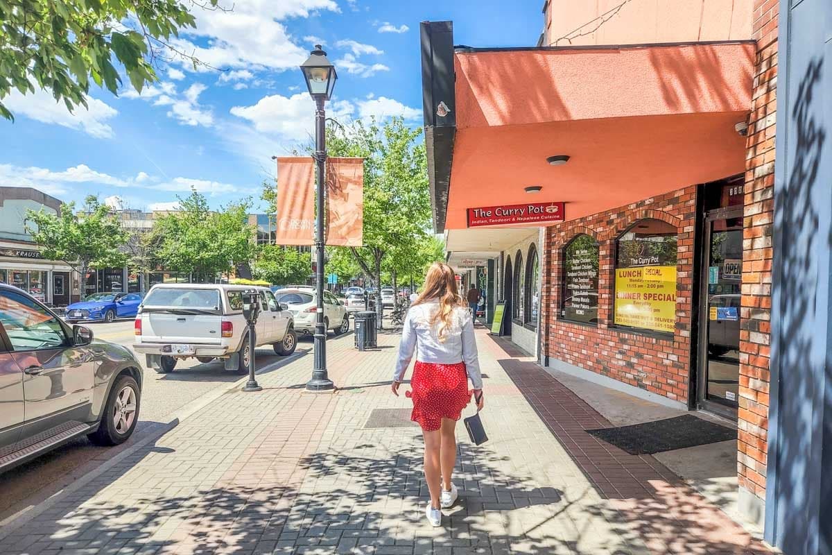 A pedestrian walks along a tree-lined downtown sidewalk past brick storefronts, with parked cars and decorative street lamps.